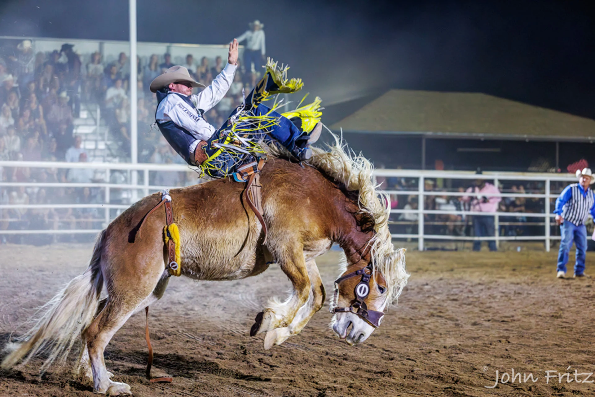 A rodeo rider in a cowboy hat rides a bucking horse in a dusty arena at night, with spectators watching from the stands in the background.
