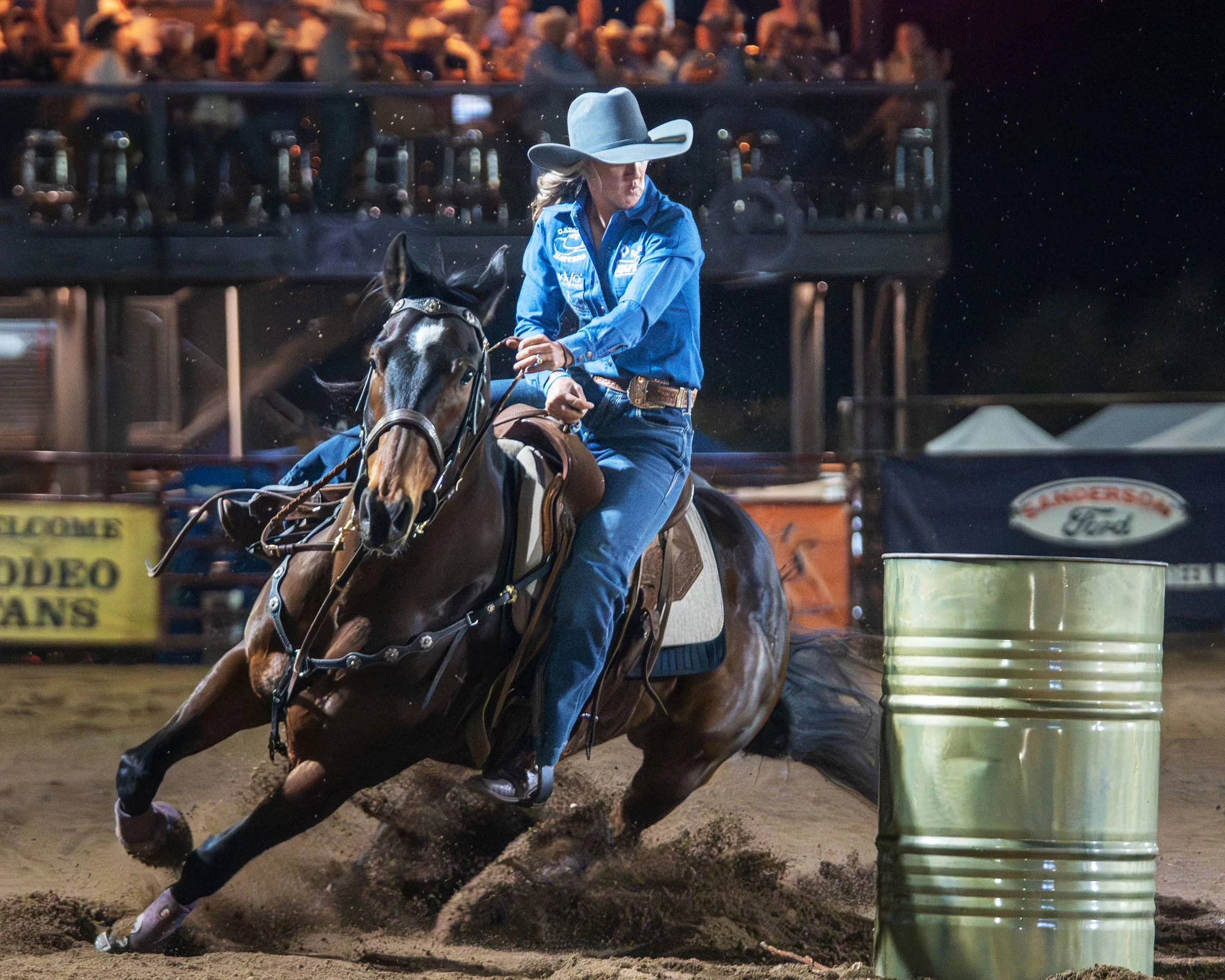 A rodeo rider in a blue shirt and cowboy hat guides a horse sharply around a barrel in a dirt arena, with spectators watching from stands in the background.