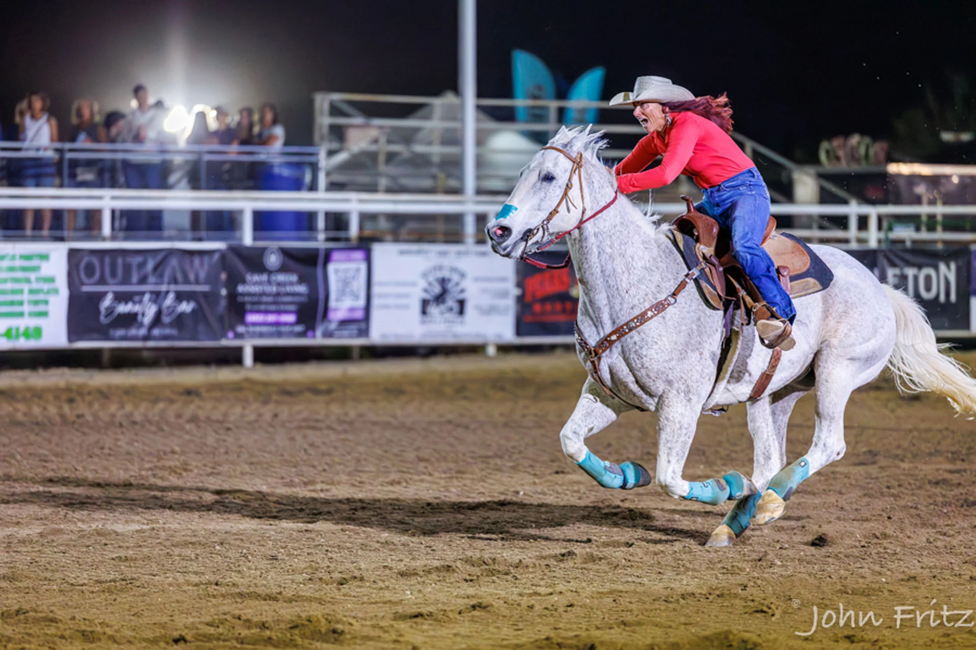 A cowgirl in a red shirt and cowboy hat rides a galloping white horse in a rodeo arena at night.