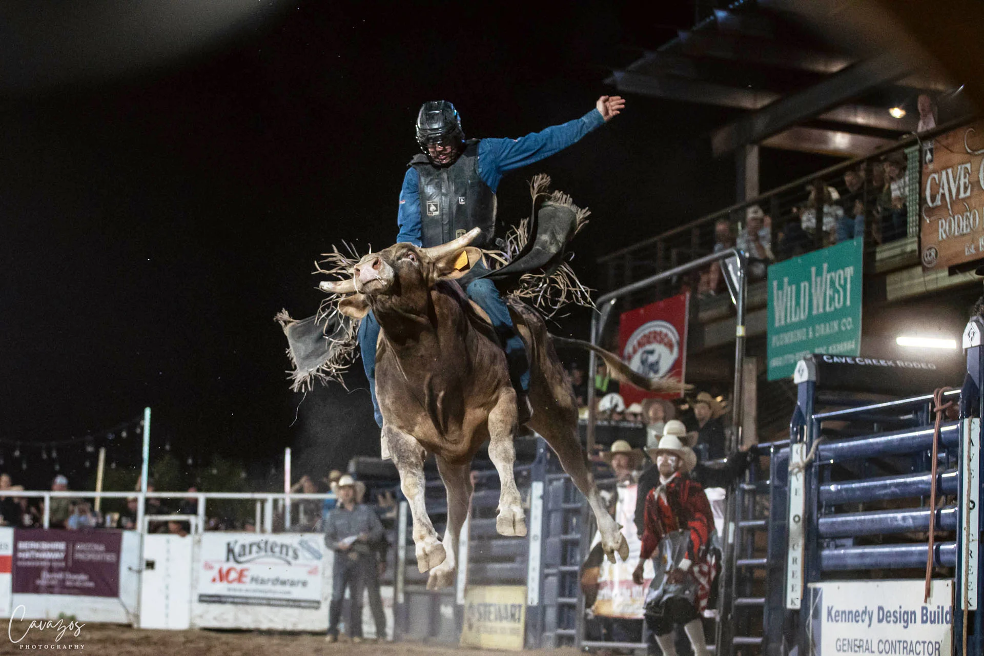 A rodeo rider wearing a helmet balances atop a bucking bull in an arena at night, with spectators, event banners, and rodeo officials visible in the background.