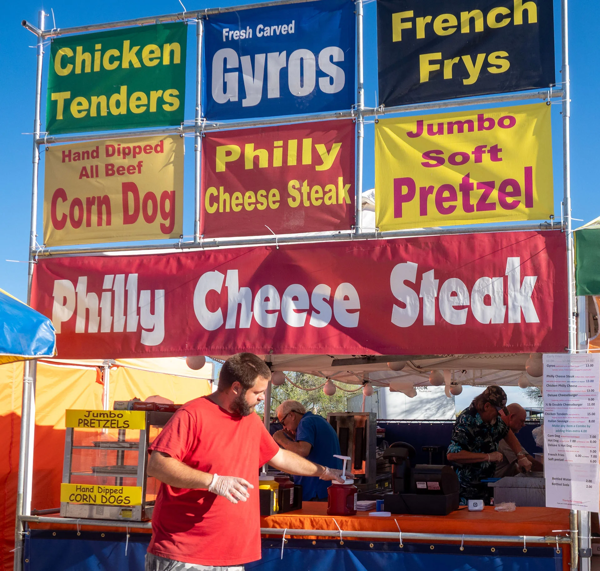 A man in a red shirt serves food at a fair booth with colorful signs advertising chicken tenders, gyros, Philly cheesesteak, French fries, pretzels, and corn dogs.