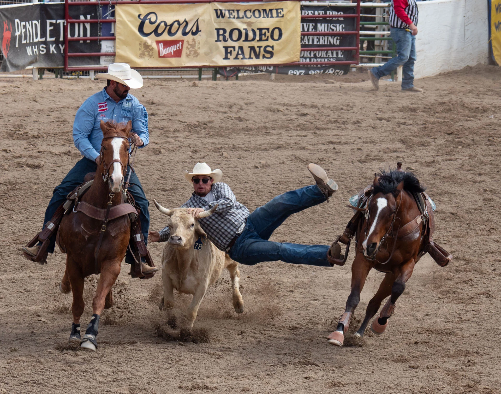 A rodeo cowboy is falling off a horse while trying to wrestle a steer, as another rider assists nearby. The scene happens in a dirt arena with rodeo signs and spectators in the background.