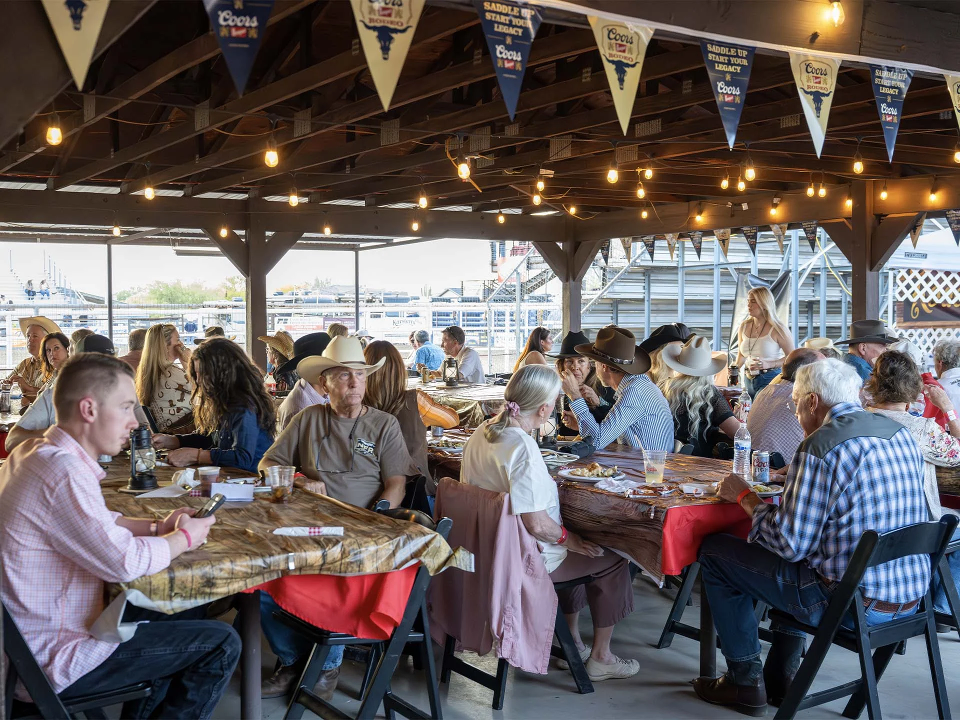 People wearing western attire sit at picnic tables under a cover at the Stockman's Club dining area, eating and socializing.